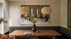 Dining nook with banquette green leather seating, a wooden table, a pair of lantern lights hanging above, an abstract painting of people dancing behind, and a large floral arrangement in a brown vase