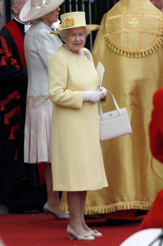 Queen Elizabeth wearing a yellow coat and an ivory purse standing on a red carpet outside Westminster Abbey on William and Kate's wedding day