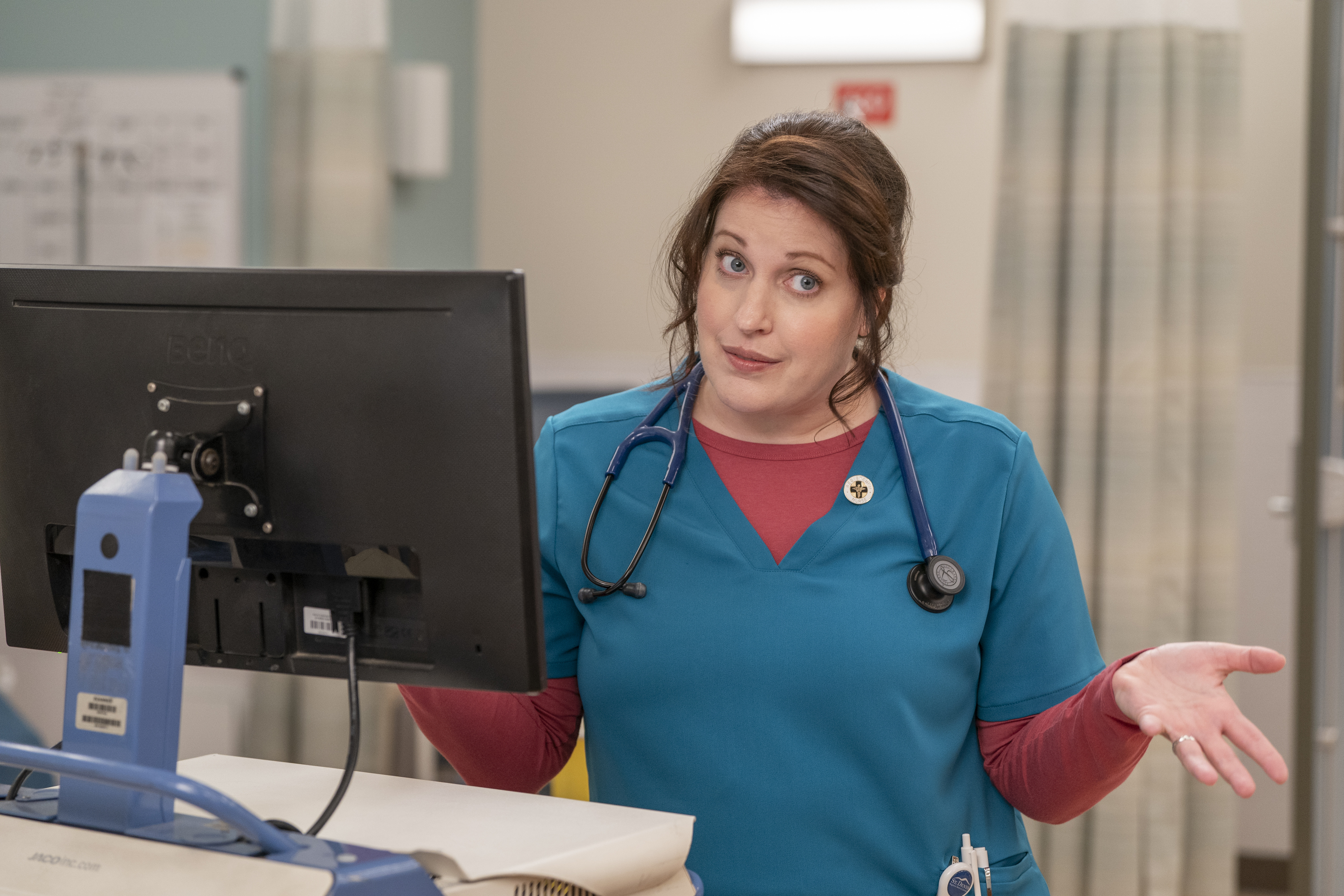 Fargo and Why Women Kill star Allison Tolman as nurse Alex wearing scrubs, standing by a computer screen and holding her hands out as if to say, 'What do you want me to do about it?'