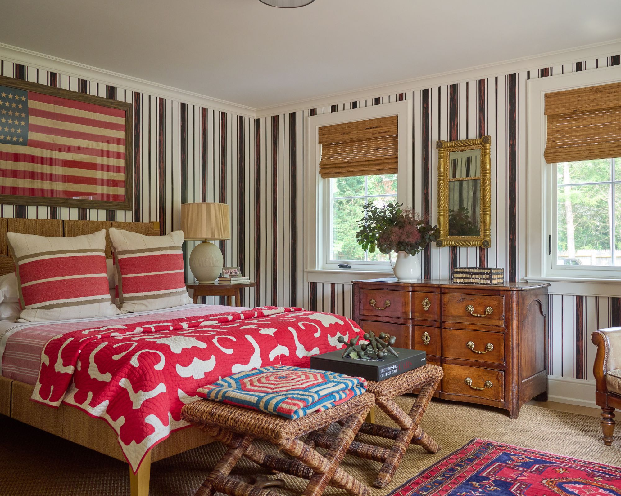 Bedroom with striped walls, an antique dresser, two rattan stools at the end of the bed, a bed with a red and white vintage quilt on top, and a vintage American flag framed above the bed.