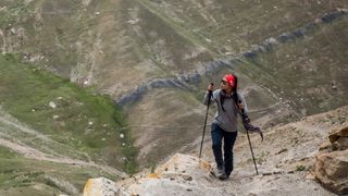 A woman wearing Montane Women's Terra Stretch Lite Trousers and using long hiking poles walks along a dusty mountainside.
