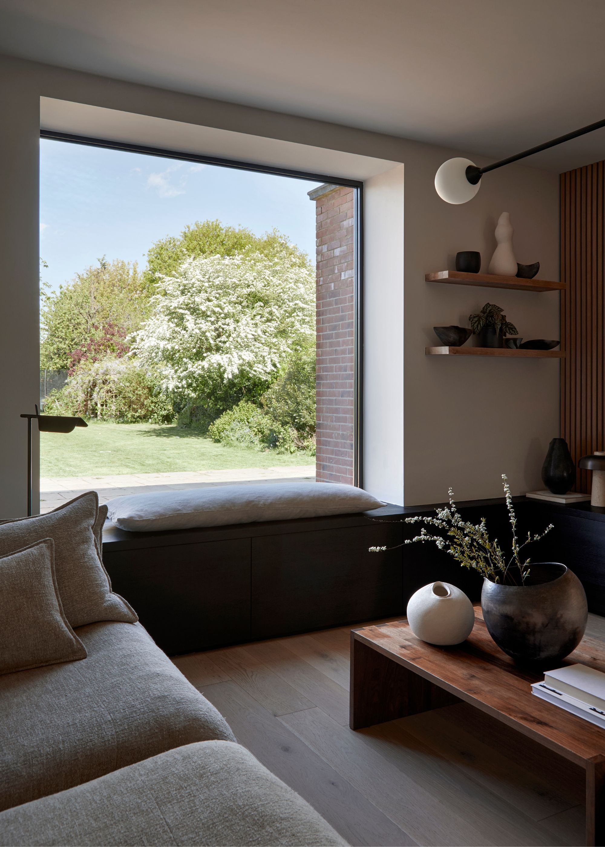 A living room with window seating looking into a modern garden. In the living room is also a large coffee table with two large vases on top of it, as well as coffee table books. Beside the window are open shelving units with decor pieces on top.