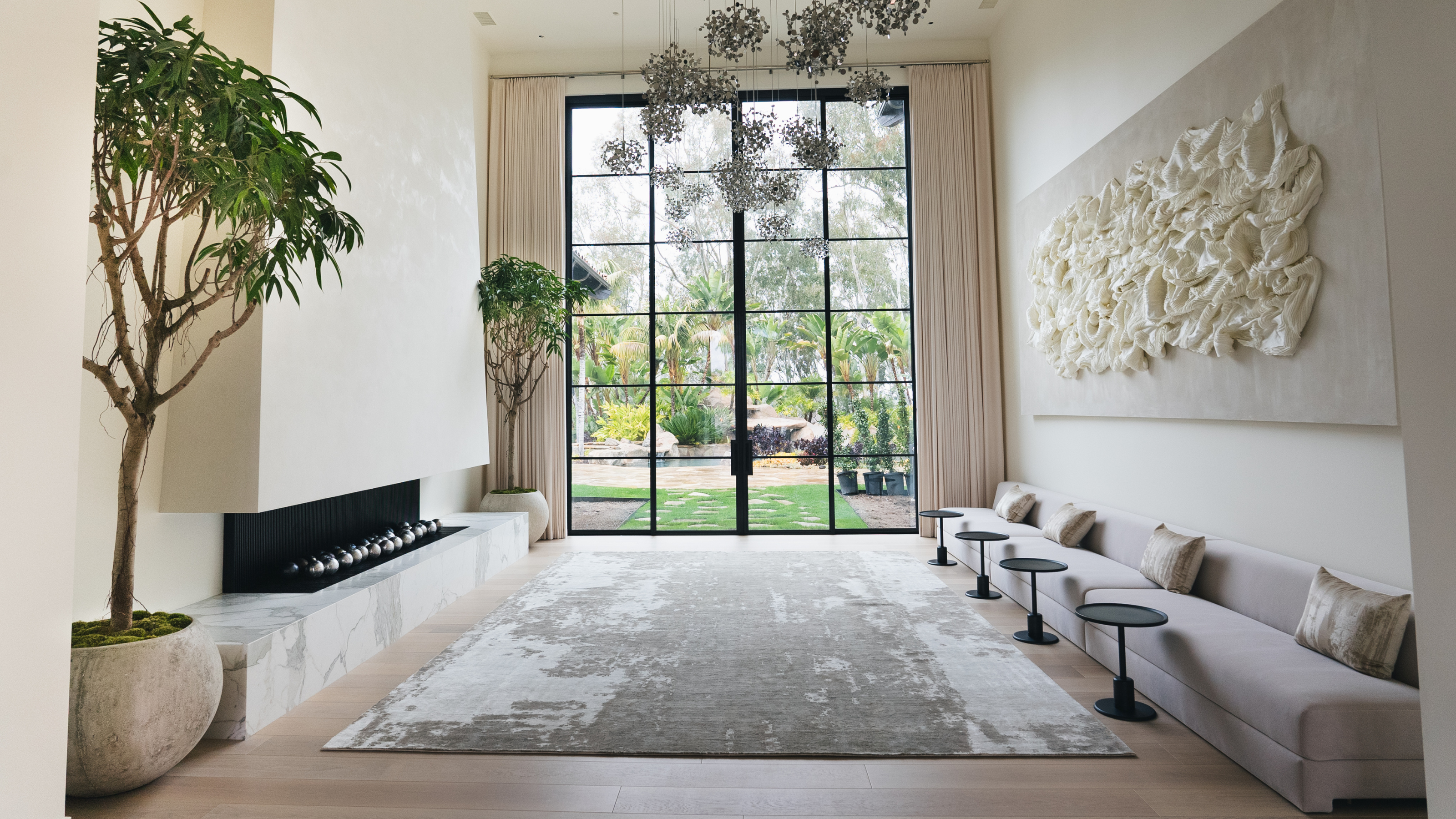 A large modern living room facing crittal style doors to the garden with a large fireplace with two large indoor potted trees either side facing banquette seating with four small round black side tables
