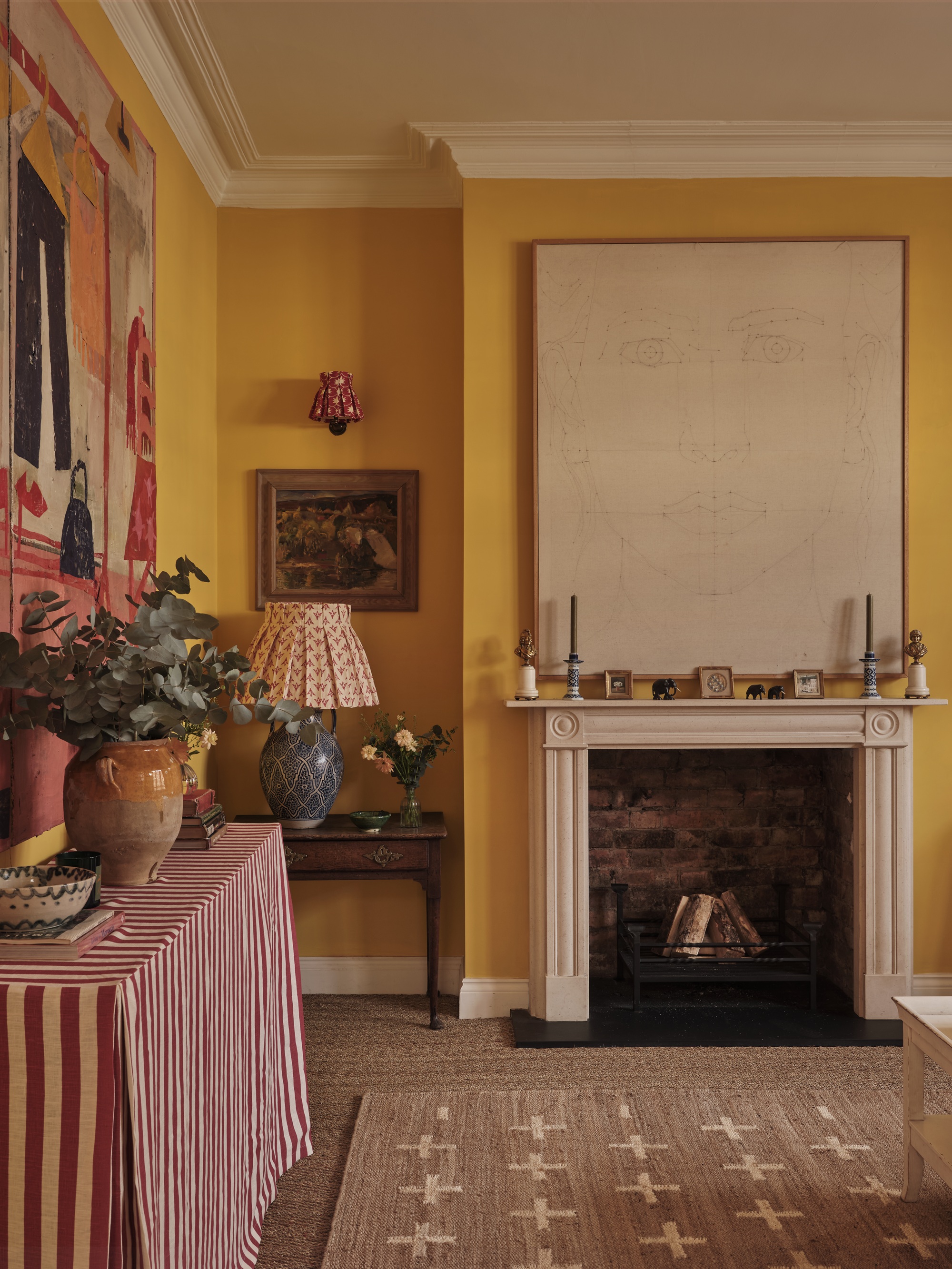 yellow living room with yellow walls, jute rug, large artwork above an original fireplace, with cornice and white ceiling, console table with red and white striped tablecloth, vase of flowers and small ceramics