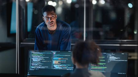 An office worker speaking to a colleague in an office at night. The co-worker is seated in front of computers