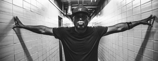 A black and white shot of Chuck D in a corridor wearing a t-shirt and baseball cap. He has his arms out touching the walls and is looking straight down the camera.