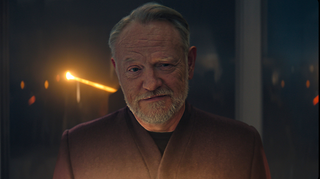 A bearded man stares at us with a piercing light in the background