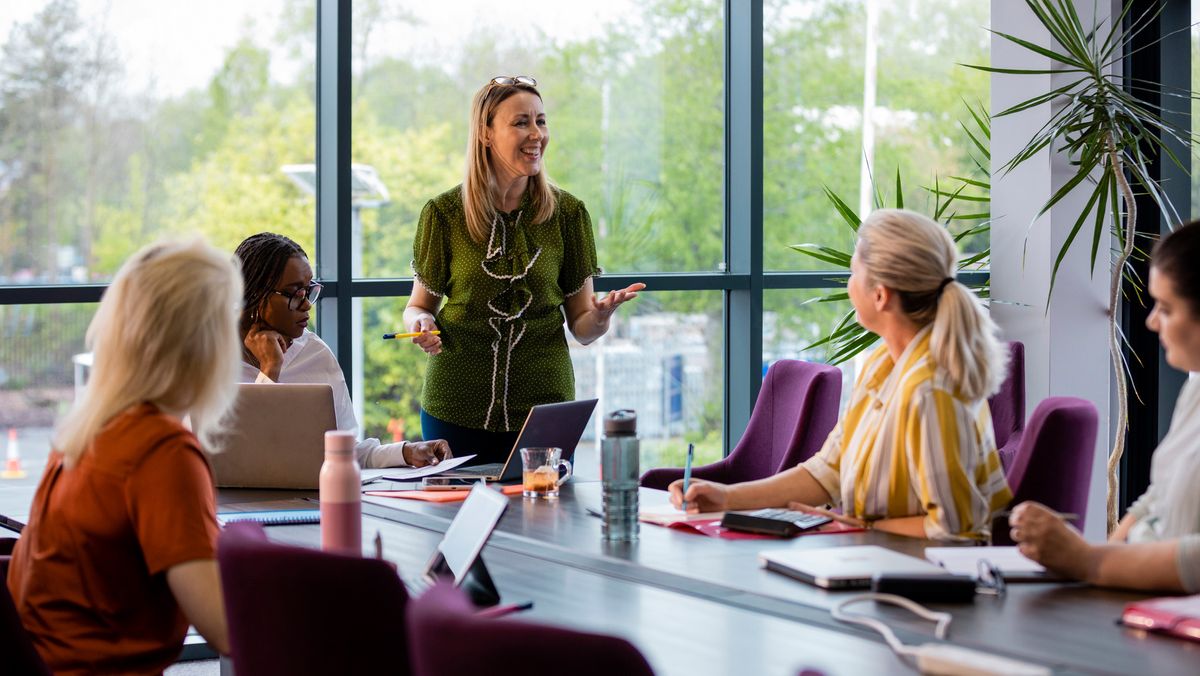 A group of women sat in a boardroom, looking at one woman who is stood at the head of the table giving a talk.