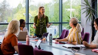 A group of women sat in a boardroom, looking at one woman who is stood at the head of the table giving a talk.