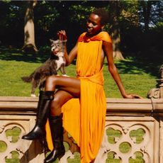 A model sitting on a stone wall wearing a draping wrap around orange dress and black boots with a small grey dog standing on the wall and her lap