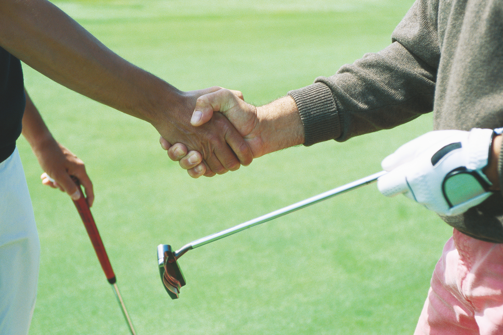 Two golfers, one wearing a glove, shaking hands while also holding their golf clubs