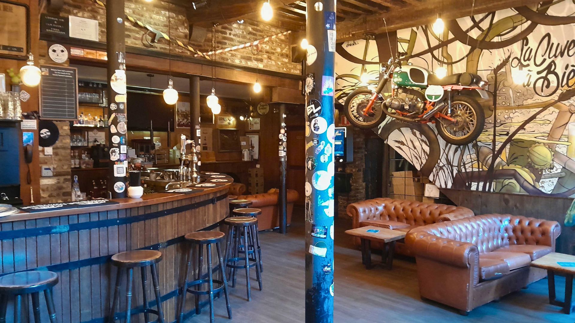 Interior of La Cuve &amp;agrave; bi&amp;egrave;re. Rustic bar interior with a wooden curved bar, high stools, leather sofas, and artistic graffiti. A vintage motorcycle hangs on exposed brick, lit by warm bulbs