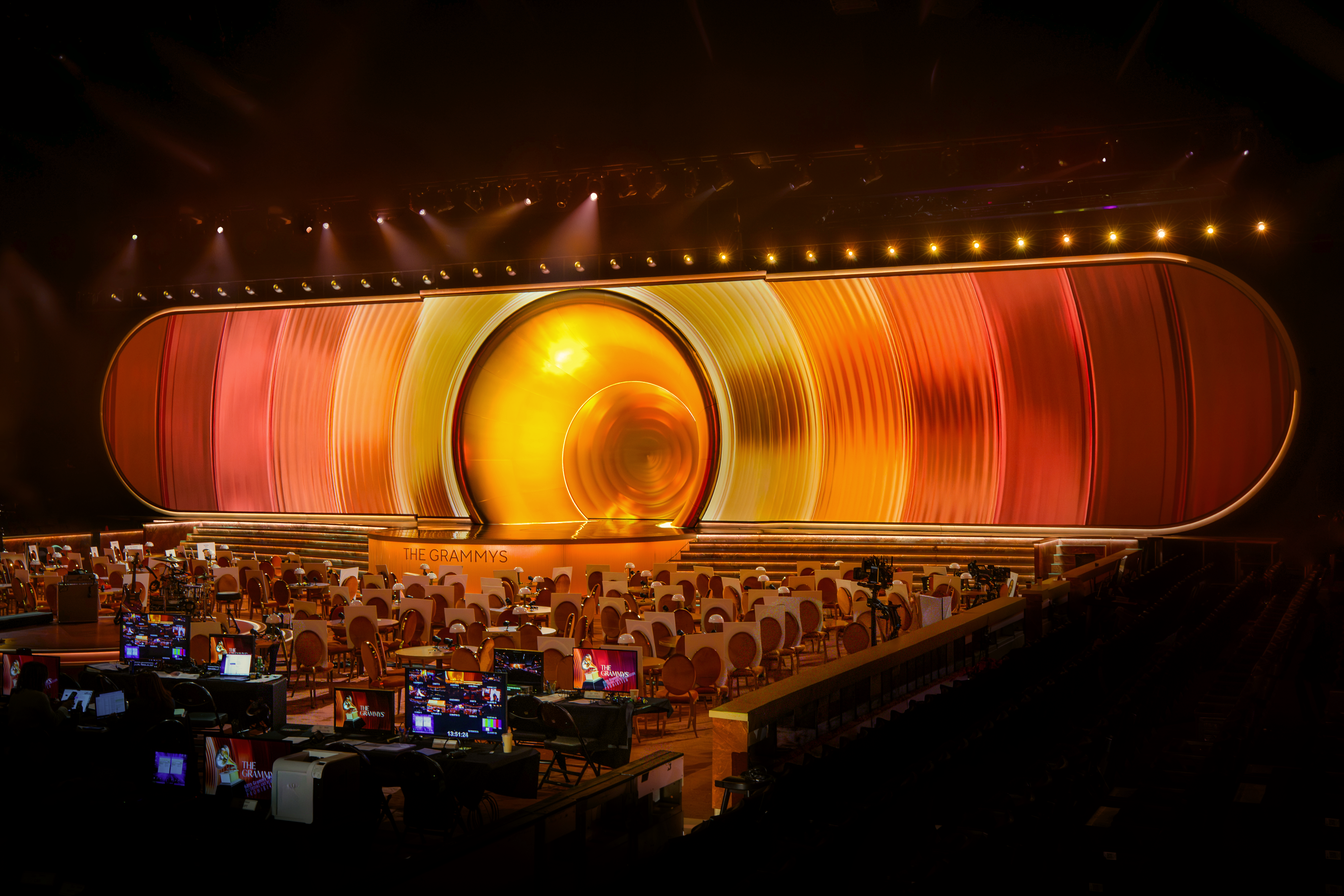 An aerial view of a performance stage with a sculptural, abstract structure of orange, red, and yellow swathes, foregrounded by a series of ceremony round tables with lamps as centerpieces.