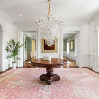 white panelled hallway with pink patterned rug, round wooden table and chandelier