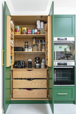 Pantry hidden within kitchen units with green exterior and wooden interior; containing appliances and produce in clear lidded jars