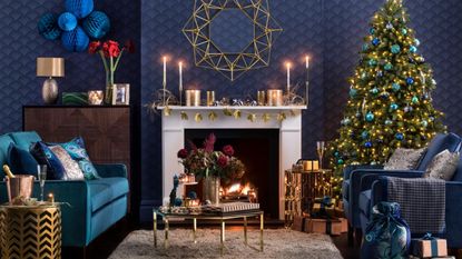 Dark blue livng room, Christmas tree with Christmas decorations, white fireplace, lit fire, gold coffee table, blue sofas with patterned cushions.