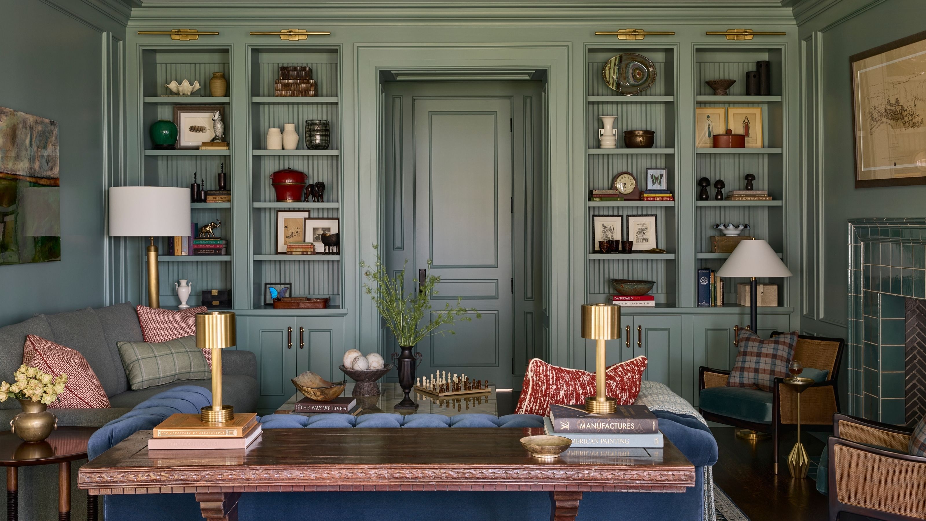 Sage green library room with floor-to-ceiling built-in bookcases surrounding a central doorway, featuring a blue tufted sofa, wooden console table, and decorative brass table lamps.