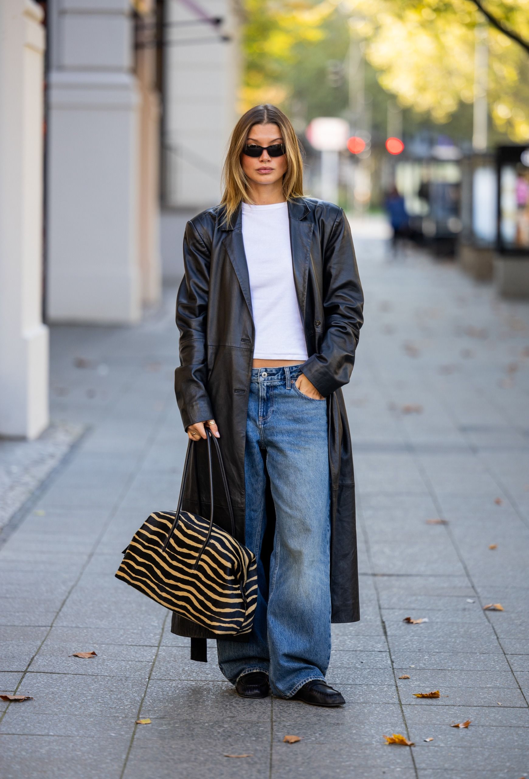 A woman stands outside wearing a white T-shirt with a black leather jacket, blue jeans and a tiger-print bag.