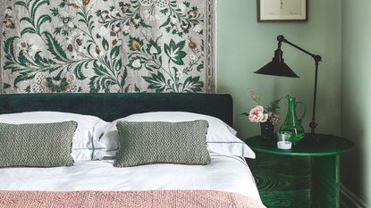 Pale green bedroom with white sheets, sage green cushions, a pale pink throw and a green and white floral wall tapestry