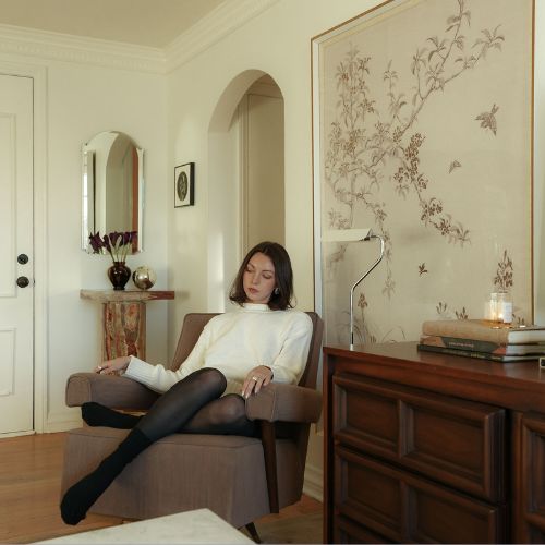 Image of a woman in a white top and black tights sitting in a brown chair in a traditional living room.
