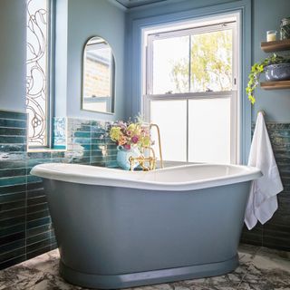 a bathroom with a large painted freestanding bathtub, marble-look flooring, tiled walls and gold taps