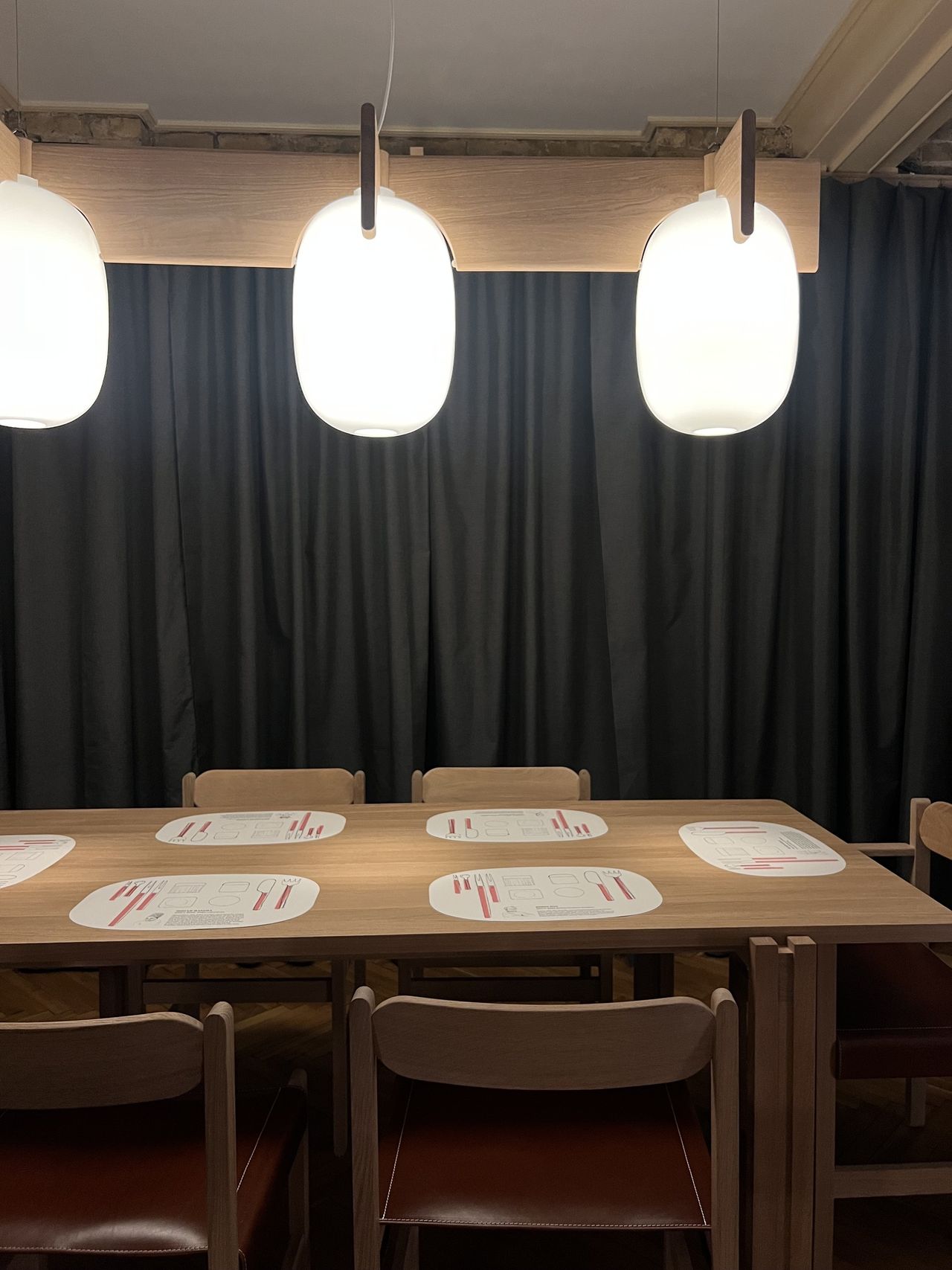 Image of a wooden table with wooden chairs with red seats around it. There are placemats on the table and a large, mid-century-style pendant over the table.