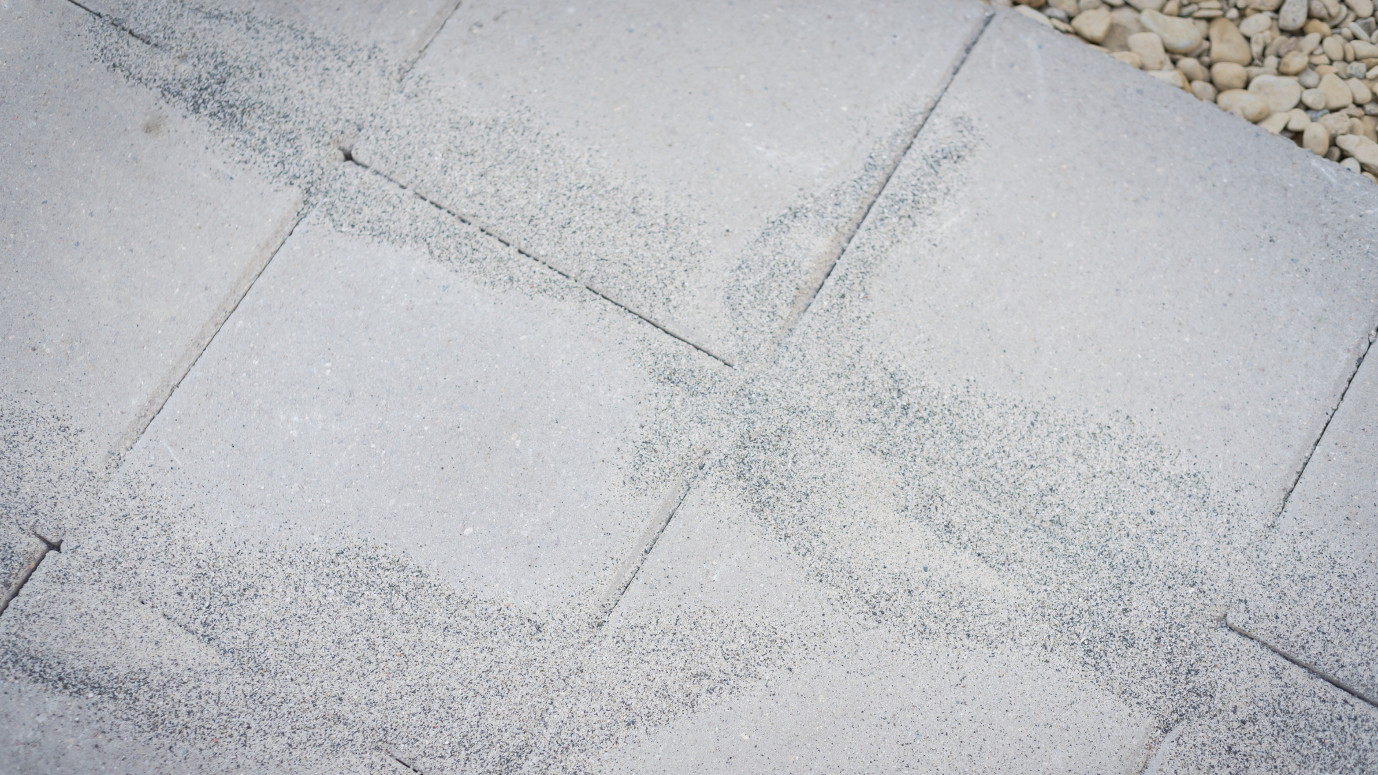 polymeric sand on grey outdoor tiles with pebbles in top right corner