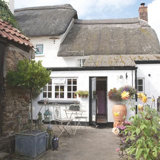 Patio area in garden with metal bistro set outside white cottage with potted bay tree and dahlias in the foreground