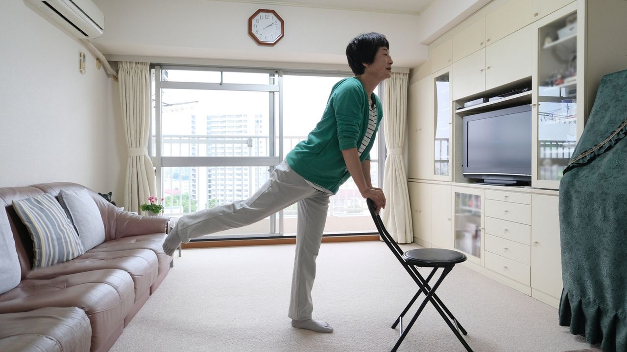 woman indoors standing on one leg while holding the back of a chair, sideways to the camera. there&#039;s a large balcony window her with a sofa on one side and a tv and storage unit on the other. 