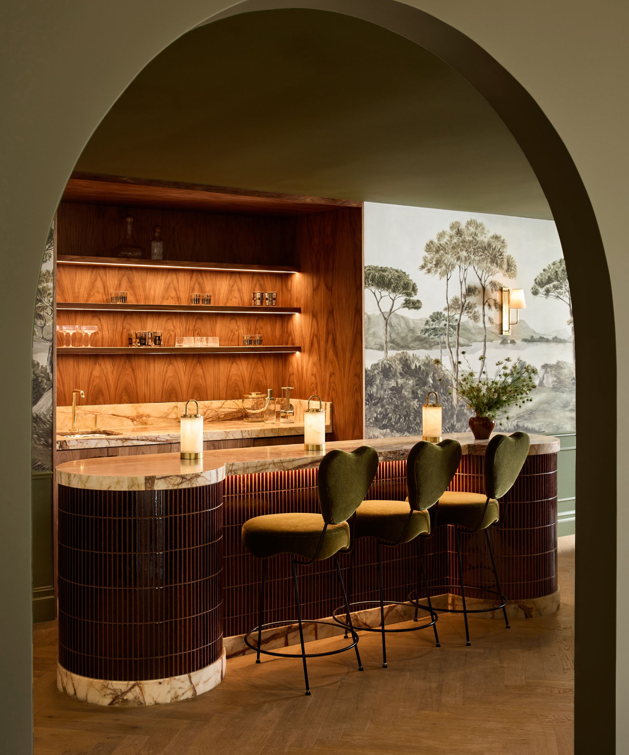 Arch doorway looking into modern kitchen with round kitchen island/bar area, three matching stools. Wooden shelving behind island, wallpaper mural to right of kitchen