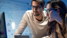 Male and female data privacy professionals looking at a desktop computer monitor in an office space, with charts and figures pictured on wall behind.