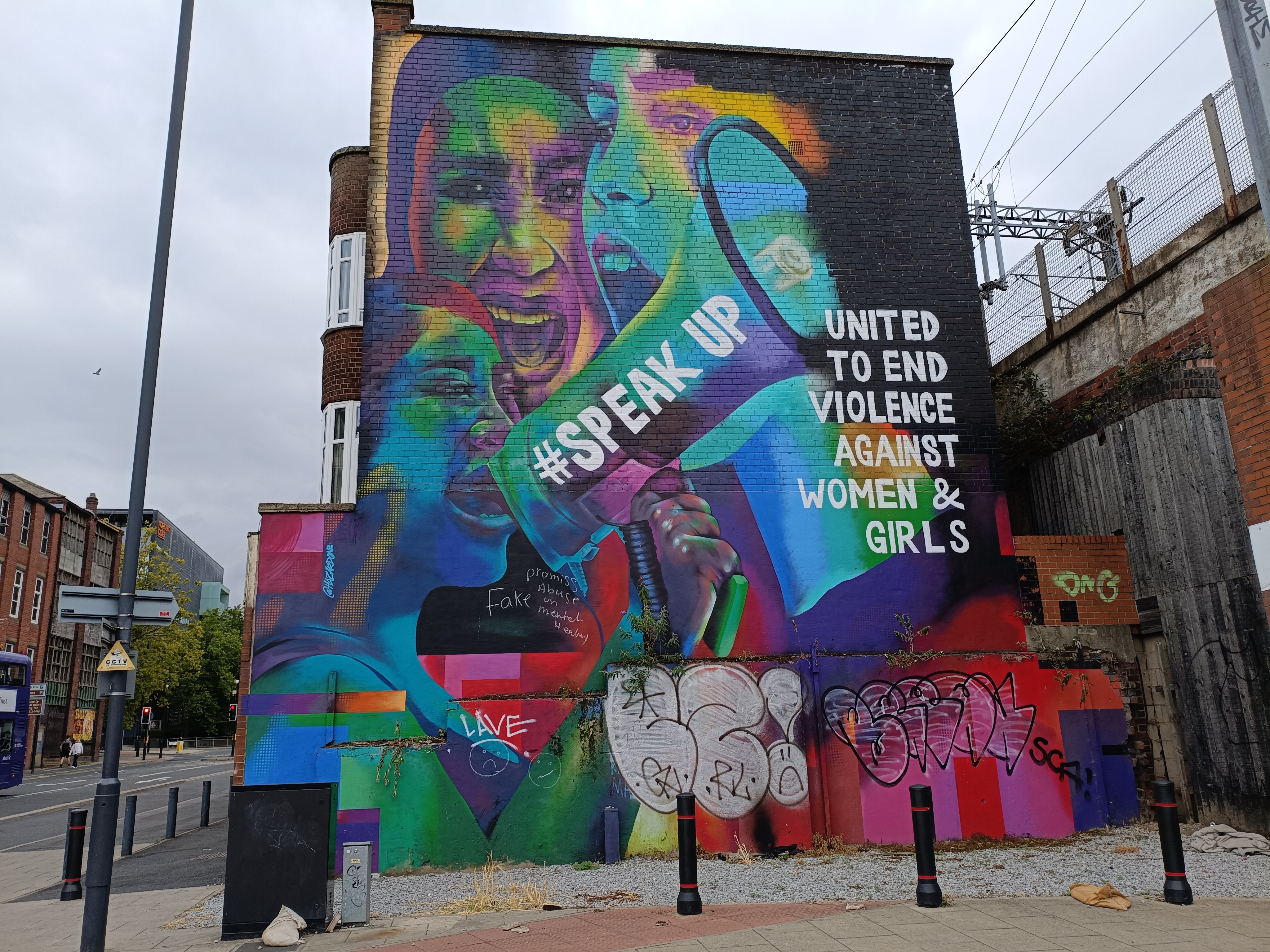 A vibrant, large-scale mural on a brick wall depicts three women in bright colors, with one holding a megaphone that says 