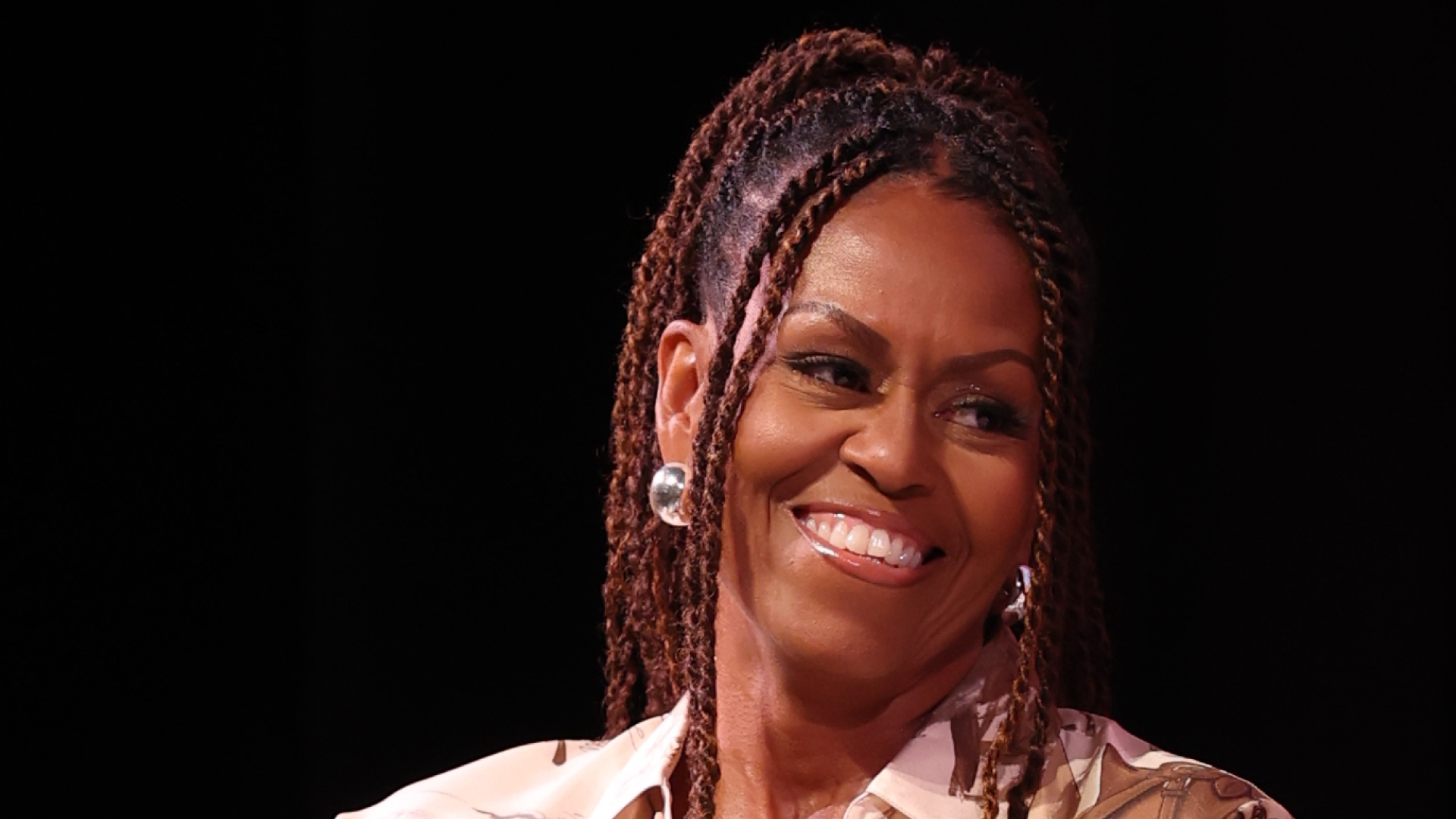 Michelle Obama with twists in her hair. She is wearing silver earrings and smiling