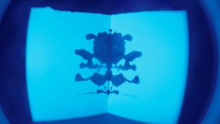 Ink blots form a Rorschach test, on a piece of folded paper lit in blue lighting and viewed through a slightly distorted lens