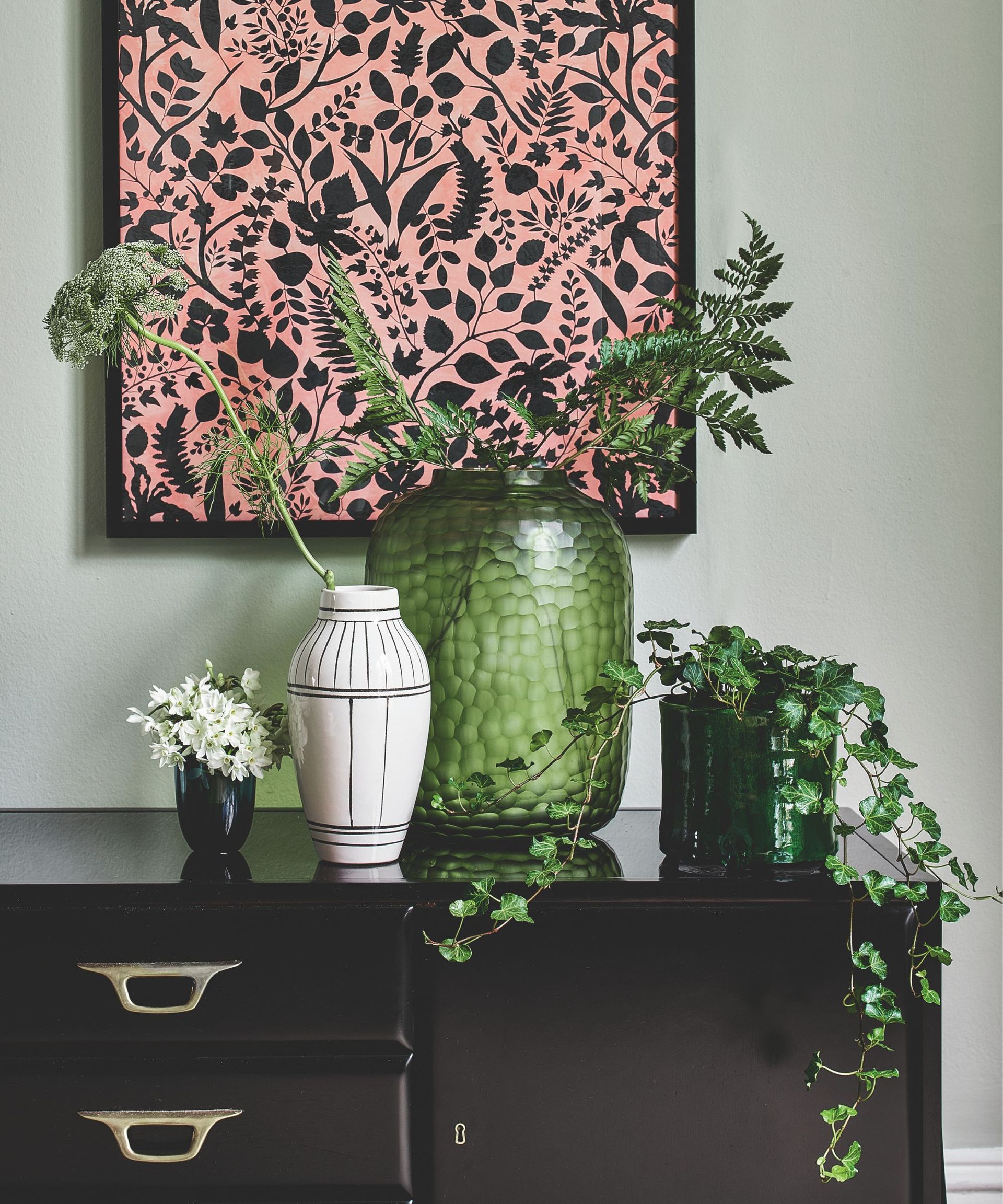 Collection of vases in green, black and white on a black sideboard, filled with green foliage ferns and ivy, with a floral pink print hung behind it