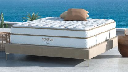 A white Saatva Classic Mattress shown on a floating bedframe against a bright blue seascape with plants, rattan tables, and neutral cushions.