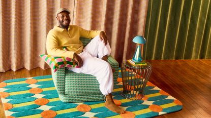 Yinka Illori sitting on a green patterned armchair on a colorful patterned rug with a side table and lamp as part of his collaboration with Dunelm