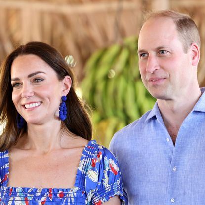 Kate Middleton wearing a blue floral dress smiling standing next to Prince William, who is wearing a blue button up, with bananas behind them