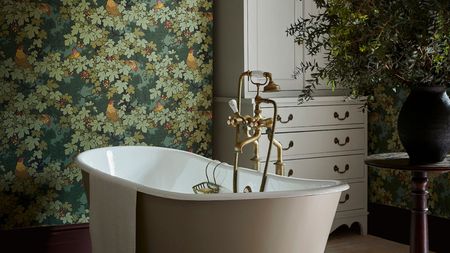 A large free-standing bath in the center of a dark, moody green bathroom. A white armoire in the background, and a side table with a large plant beside it.