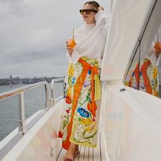 ali poses on a boat with a drink in hand wearing a white top and a colorful skirt
