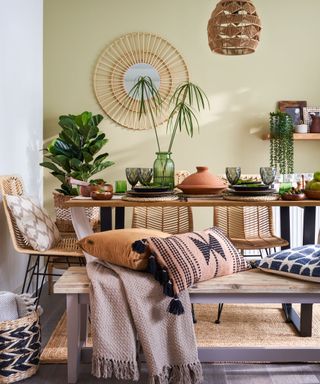 A dining room with a green wall with a rattan mirror, a dining table bench with gray glassware and plants, a wooden bench with gray and orange throws and pillows, and a jute rug on a gray wooden floor