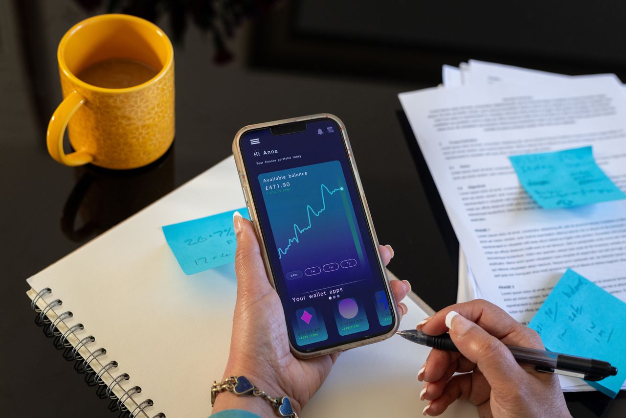 Close-up shot of a smartphone being held in a woman&#039;s hand as she takes notes. The smartphone has a fintech application open on it showing figures and data.