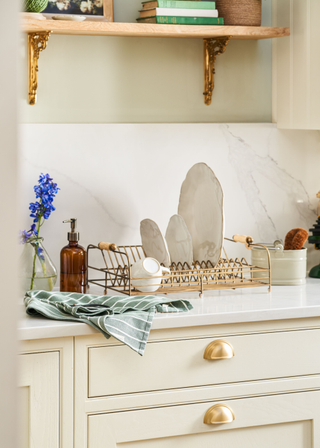 Image of an off-white kitchen with white marble backsplash and counters. There is a dish drying rack and a glass, amber soap dispenser on the counter.