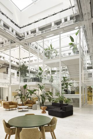Seating and open atrium at Sid Lee’s international hq, Tall potted plants, round table and wooden chairs, black corner sofa and brown leather armchairs, white stone floor, high white glazed framework and stairwell, high white ceiling with skylight