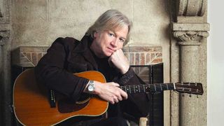 Justin Hayward sitting in front of a fireplace holding an acoustic guitar