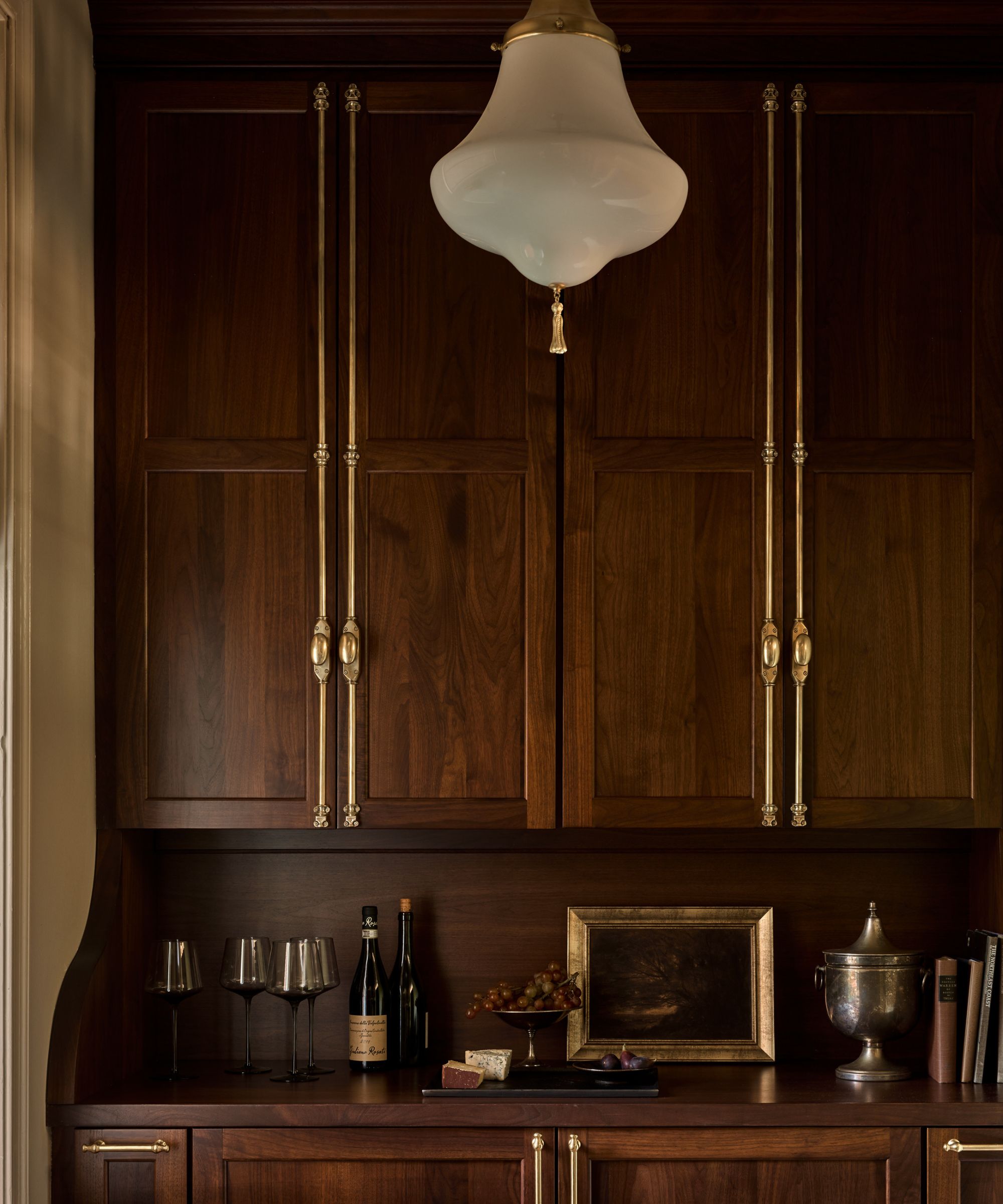 Bar pantry with dark wooden cabinets and cupboards, gold joinery and hardware, a vintage glass pendant light, barware on the countertop, a vintage painting, and an antique silver ice bucket.