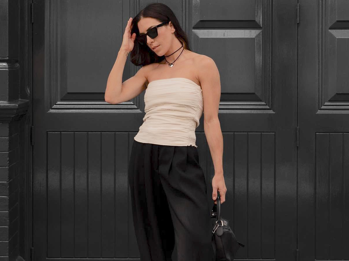 influencer Jessica Skye poses in front of a black wall in London wearing black sunglasses, a black cord necklace with a silver pendant, a beige ruched sleeveless top, black trousers, and a black bag