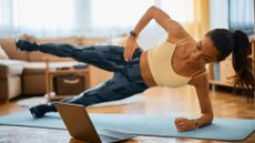 woman performing a side plank with a leg lift on an exercise mat in a home setting, looking down at a laptop on the floor.