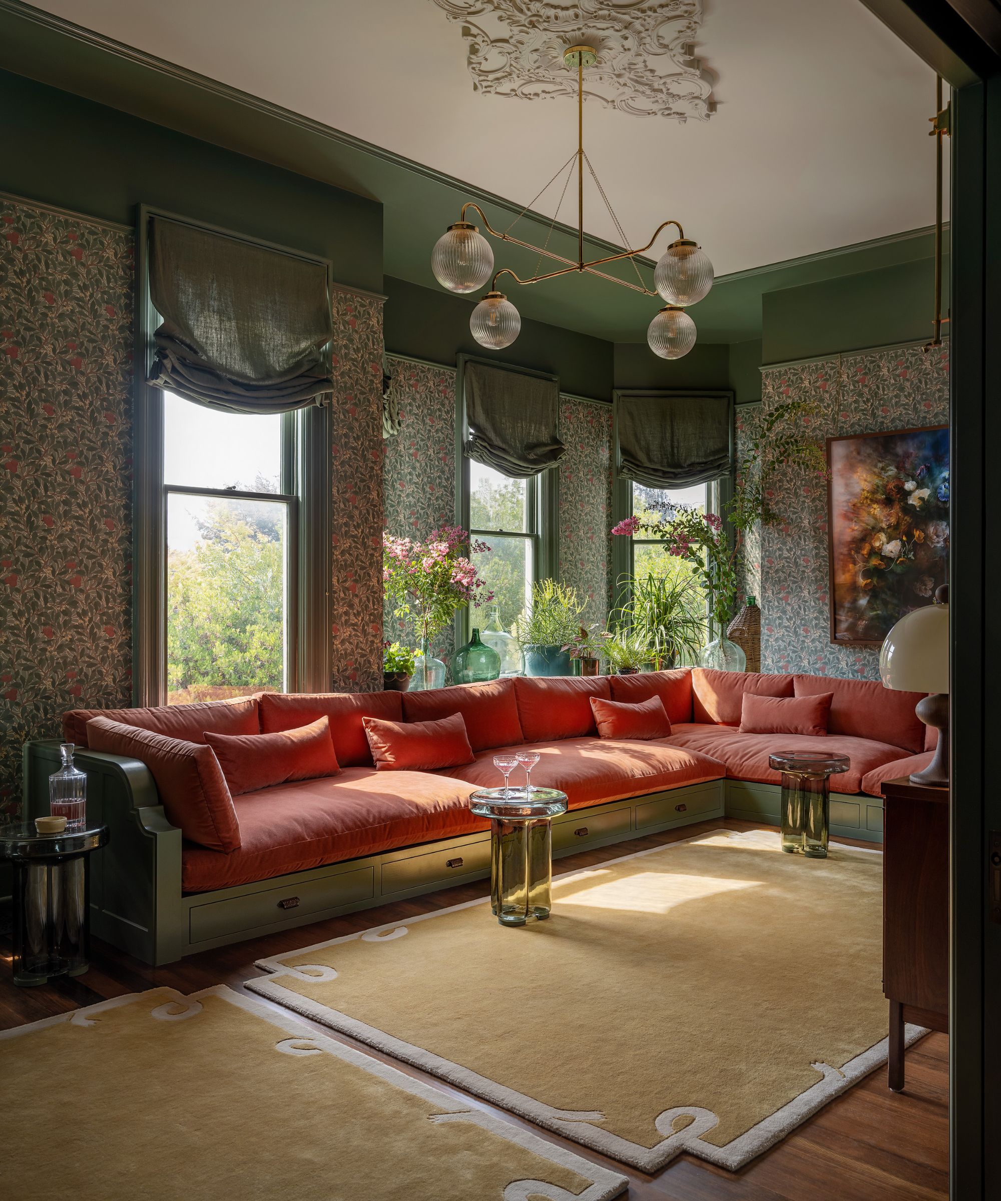 A living room in a Victorian home with green patterned wallpaper, dark sage green detailing, a large coral corner sofa, three tall windows with green relaxed Roman blinds, and two sand-colored rugs.