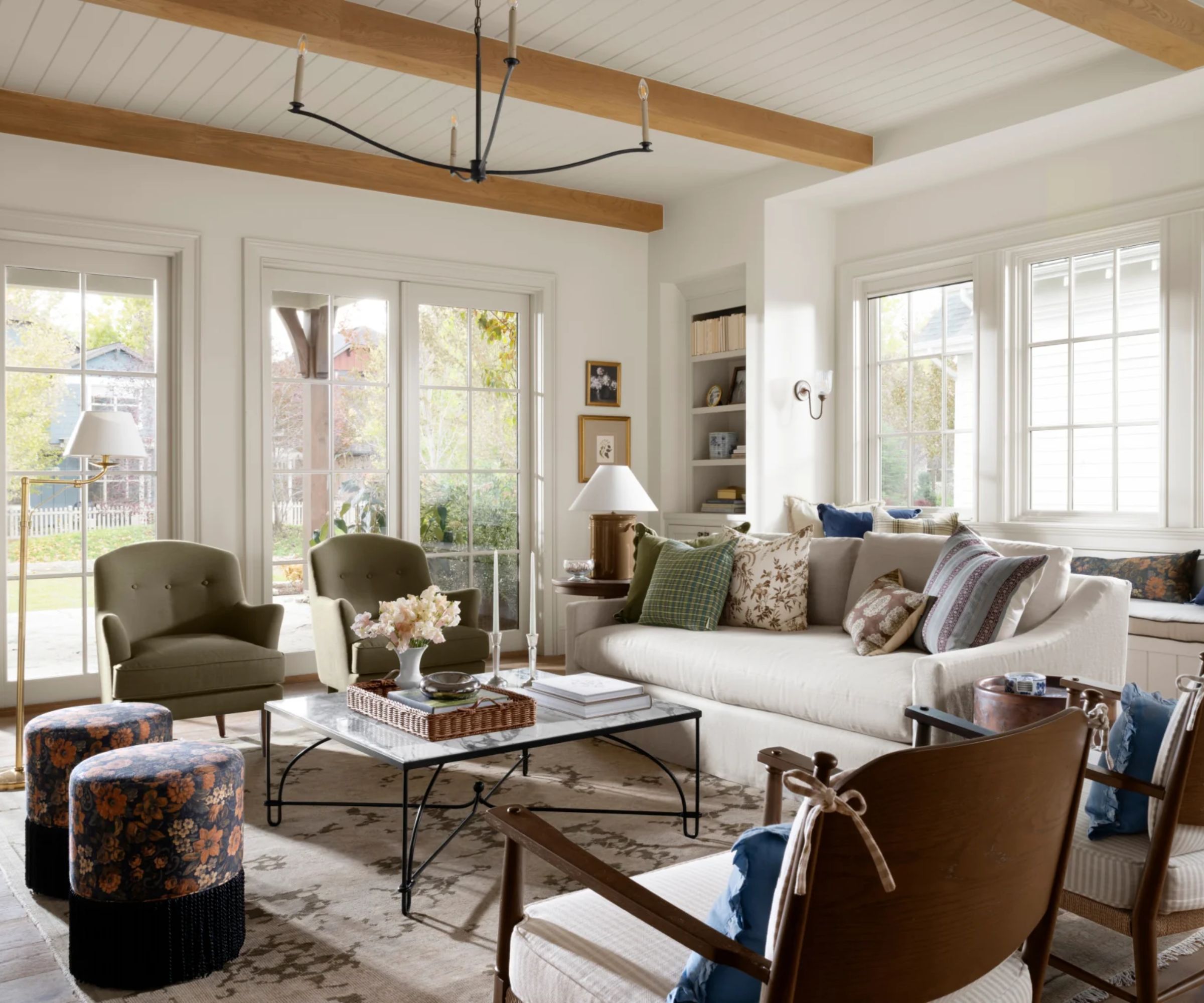 McGee and Co neutral living room with large windows and patio doors. a white couch is paired with a marble and metal coffee table with four chairs facing it in two different styles, and two navy floral fringed ottomans